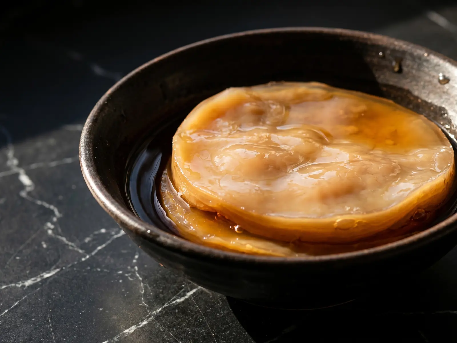 Bol de th&eacute; sombre avec un disque de SCOBY beige sur marbre noir, &eacute;clair&eacute; par une lumi&egrave;re naturelle.