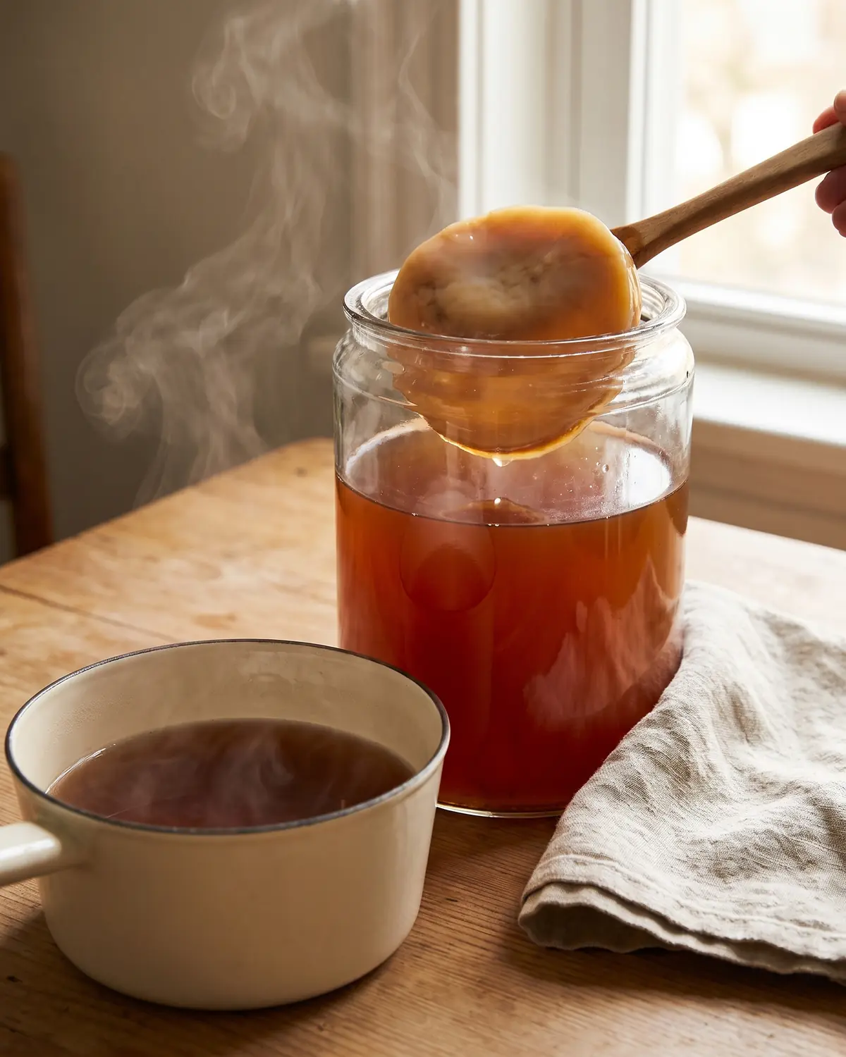 propriété kombucha Un fin nuage de vapeur s’élève d’un thé en casserole émaillée, avec un bocal de SCOBY posé à côté.