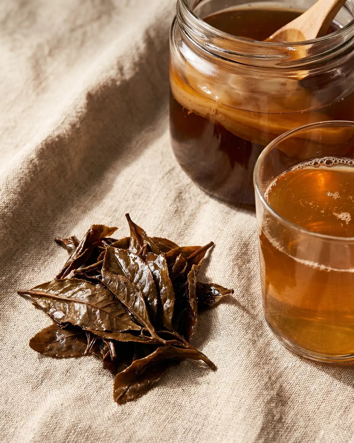 kombucha et constipation Hojas de té humides près d’un verre de kombucha sur du lin brut, dans une ambiance naturelle.