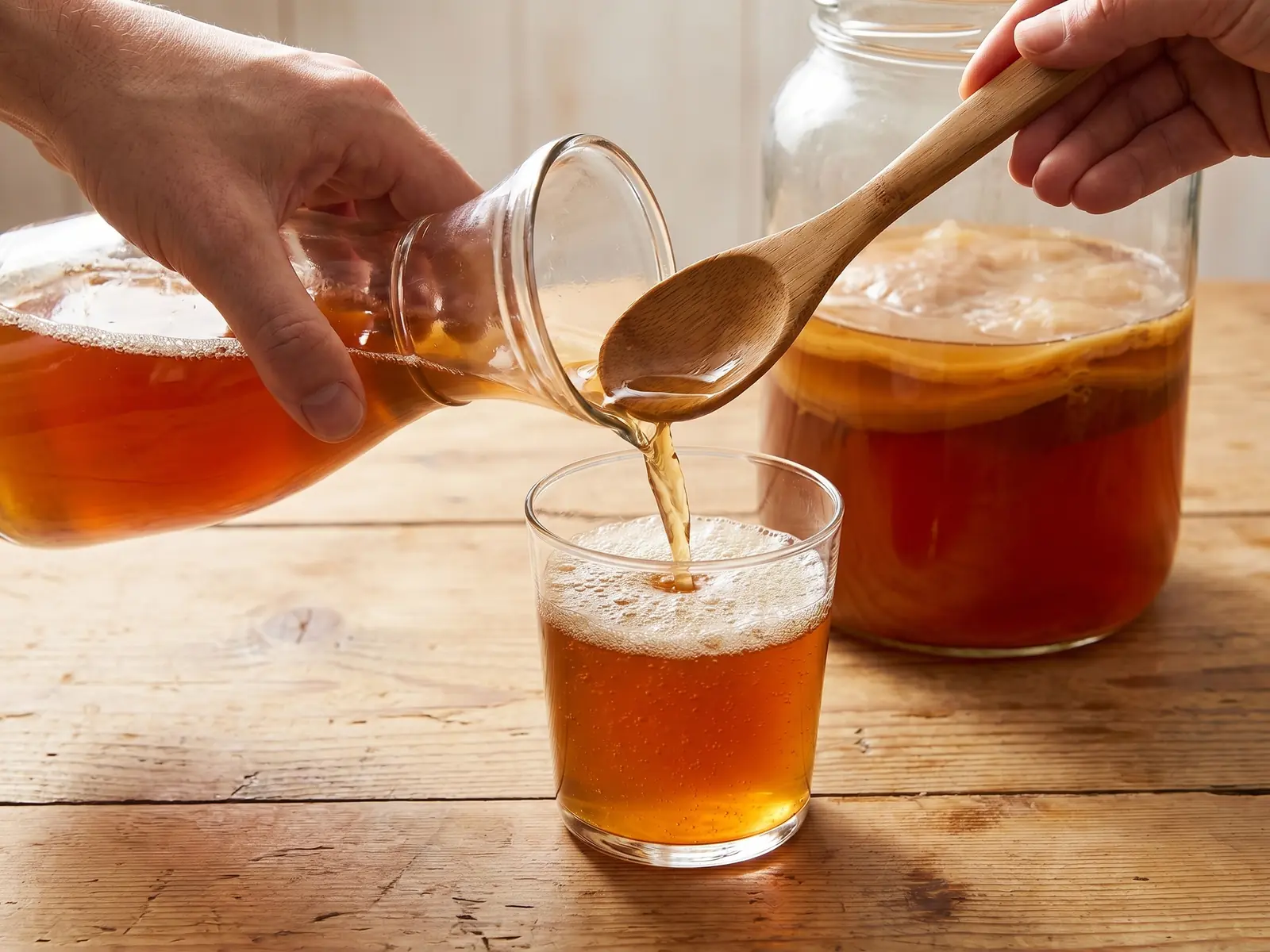Main versant du kombucha ambr&eacute; dans un verre, avec une fine mousse sur une table en pin.