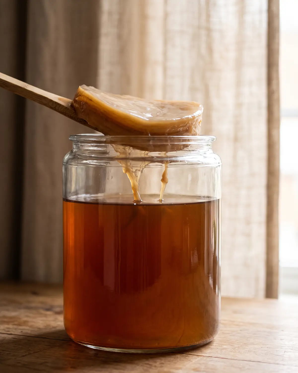 Bocal de kombucha avec SCOBY et filaments visibles pr&egrave;s d&rsquo;un rideau de lin flou.