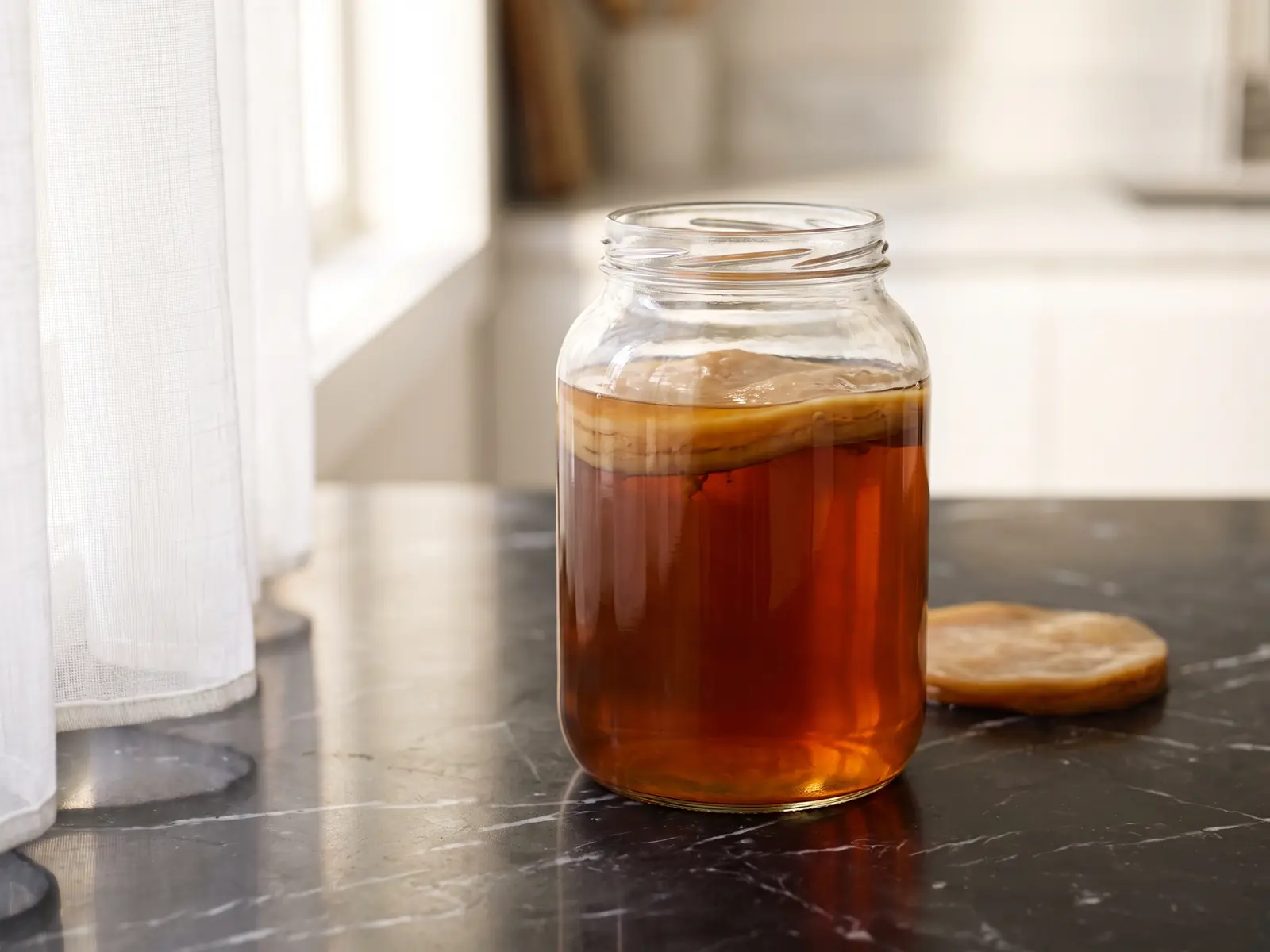 Bocal de kombucha ambr&eacute; avec SCOBY suspendu, pos&eacute; sur marbre noir avec reflet de rideau blanc.