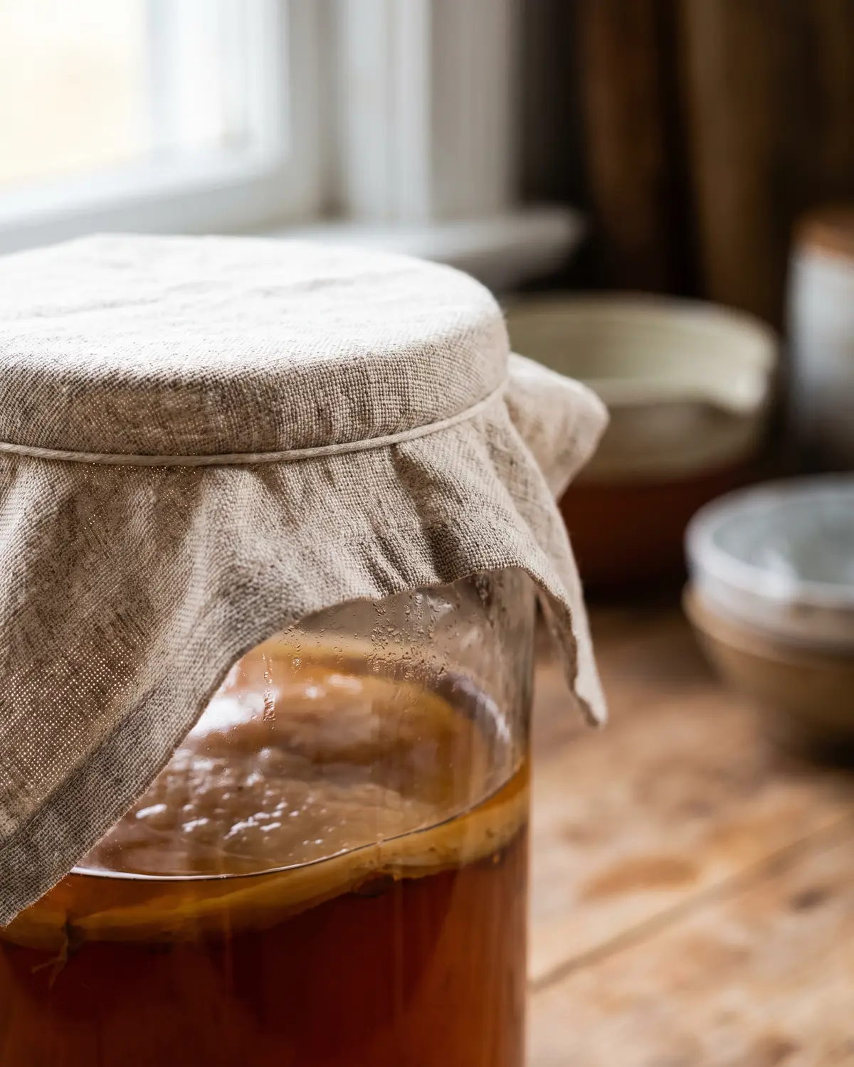 Gros plan sur un bocal de kombucha couvert de lin, avec le SCOBY flou sous un th&eacute; ambr&eacute;.